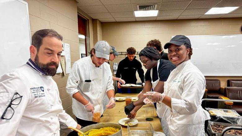 Six students and an instructor wearing chef coats spoon polenta onto plates